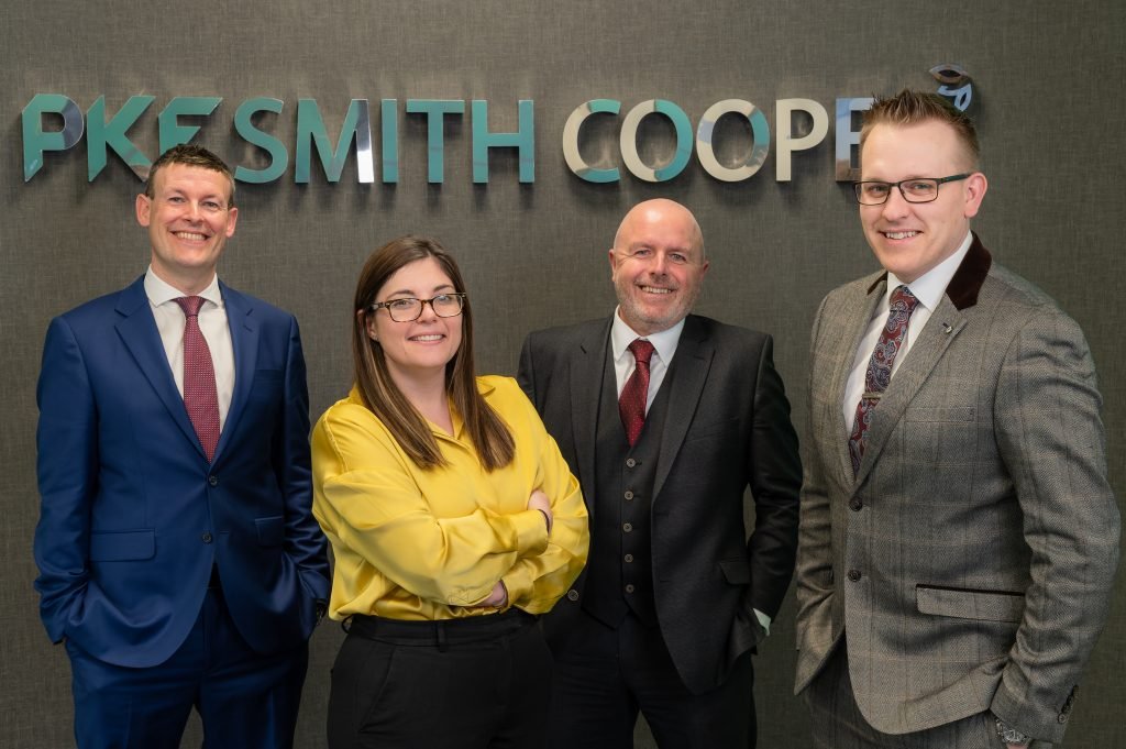 Three men and a woman dressed in professional attire stand in front of a sign that says 'PKF Smith Cooper'. They are all smiling.