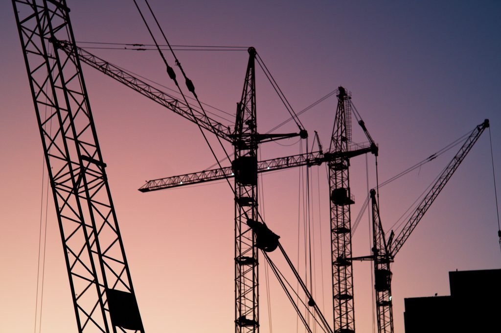 A row of construction cranes photographed with the sky as a backdrop. The sky is a mixture of purple, pink and yellow.