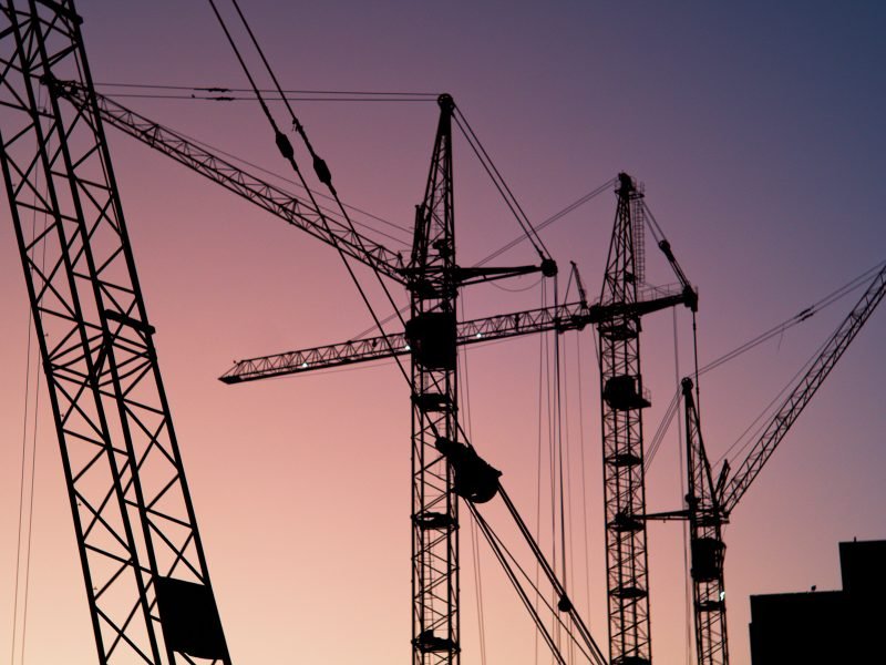 A row of construction cranes photographed with the sky as a backdrop. The sky is a mixture of purple, pink and yellow.