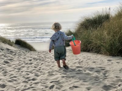 A toddler is walking on the beach towards the sea. The child's back is to the camera. The child is holding an orange bucket.